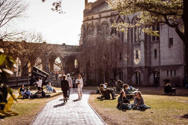 Photo of students walking and sitting in the grass and lawn chairs on BU Beach, the back corner of Marsh Chapel is seen. It is dusk and the sky is grayish white. Some of the trees have started to bloom.