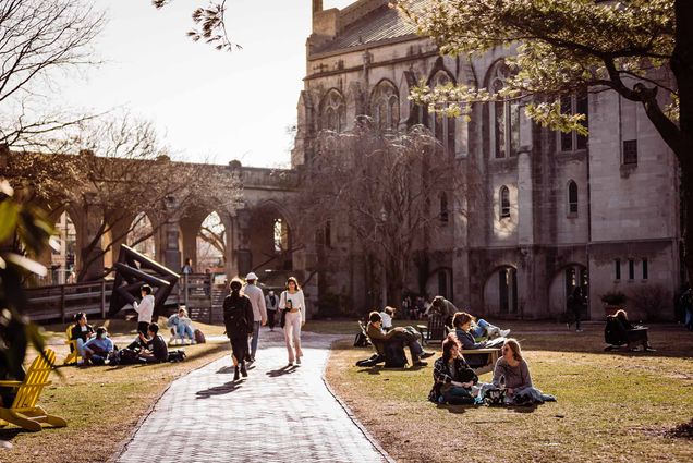 Photo of students walking and sitting in the grass and lawn chairs on BU Beach, the back corner of Marsh Chapel is seen. It is dusk and the sky is grayish white. Some of the trees have started to bloom.