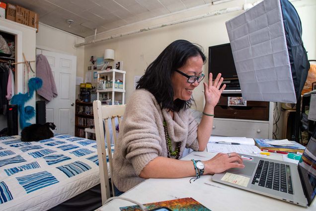 Photo of Puppeteer Leigh Baltzer, owner of Through Me To You Puppetry, smiling and waving at her computer as she attends a BU business course virtually from her Brookline home Feb 28. Baltzer is a middle-aged Asian American woman with shoulder length black hair, glasses, and she wears a tan sweater and series of chunky necklaces and bracelets.