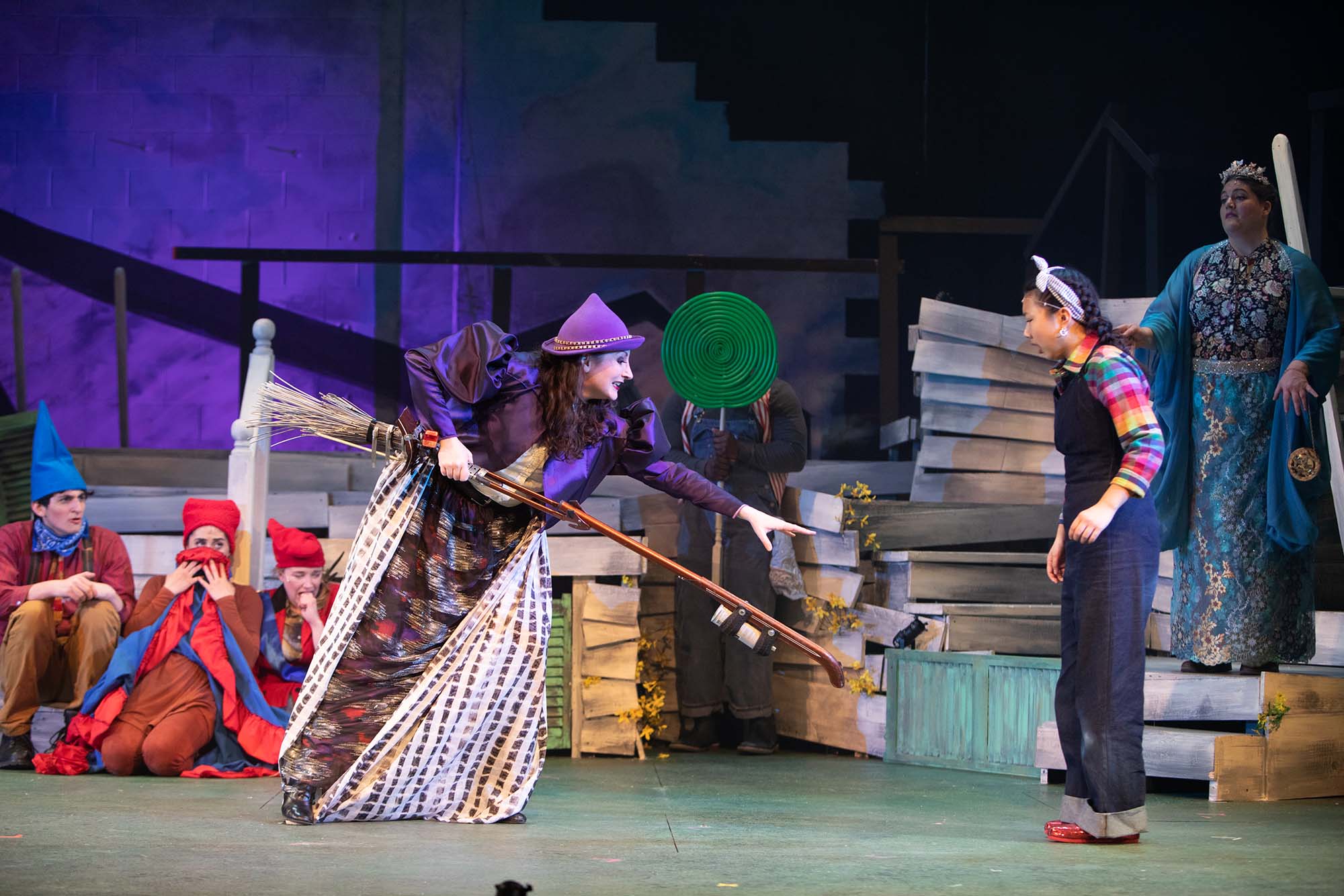 Photo of Katie Anne Clark (left) as the Wicked Witch of the West with Lily Park as Dorothy in Boston University’s production of “The Wizard of Oz,” during a dress rehearsal on Wednesday, April 6, 2022. Clark wears a purple witch's hat and long patterned dress. Park wears a handkerchief around her head and overalls. Behind them are stacked metal beams and other debris.