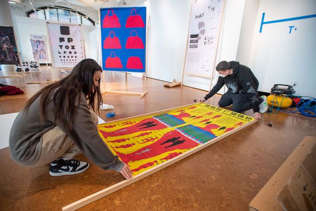 Photo of Chen Luo (MFA’22), left, helping associate professor Christopher Sleboda secure Luo’s piece, Radio Exercise, to a wooden frame for the MFA Graphic Design Thesis show April 7 in Stone Gallery. Luo has long brown hair and wears a gray sweatshirt and nike sneakers, Sleboda wears a black sweatshirt, a face mask and kneels opposite Luo. The piece features figures in red, with smaller black figures inside them, surrounded by repeated black text on a yellow background. Other pieces from the exhibition, including a large blue piece with a repeated handbag pattern in pink, are seen behind them.