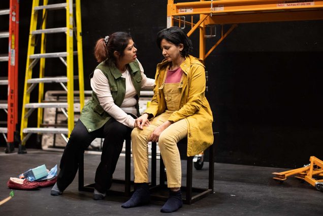 Photo of Mita Das, (left), a Bentley University Senior Lecturer of Mathematical Sciences and Prreeti Tiwari, right of South Windsor, CT, rehearsing for the Playwright's Theatre marathon. Das wears a green vest, white long sleeve, black pants and wears no shoes. She is a brown woman with brown hair. She sits on a stool and leans over to Tiwari, putting her hand on her knee. Tiwari, a brown woman with short black hair, looks forlorn, and wears bright yellow overalls.
