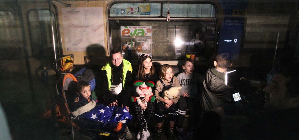 Photo of children, of various ages, sitting in a train car in a metro station that serves as a bomb shelter. They have blankets and some hold stuffed animals. Above them you can see subway advertisements. The photo was taken through a subway car window.