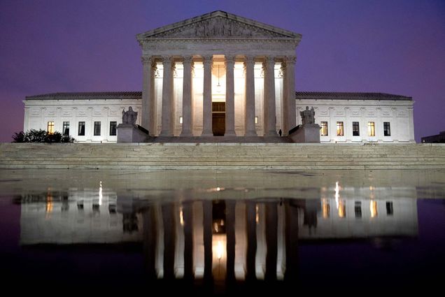 Photo of the facade of the supreme court at dusk. The white, Grecian facade is reflected in a puddle in front of the main steps. The sky is blueish purple and lights shine from behind the columns.