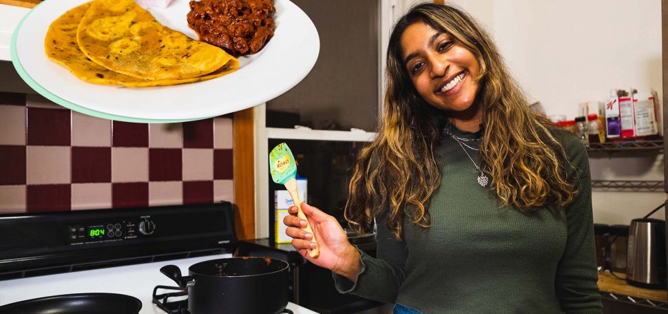 Photo of Neytra, a young Indian American woman with light brown skin and long brown, highlighted hair. She wears a green sweater and holds a spatula in her right hand. Behind her, you can see two black pots on a stove and a kitchen backsplash. At the top left of the photo is an image of the dish she cooked: curry made with red kidney beans and pureed tomatoes and flavored with spices like cloves, tumeric, and cinnamon with a side of roti. White play button is overlaid.