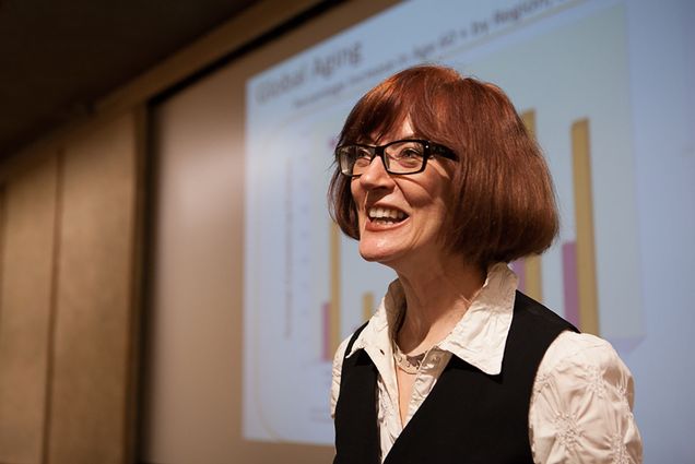 Photo of Valerie Fletcher, a middle-aged white woman with short red hair and bands. She smiles and wears thick black glasses and a white and black collared shirt. Behind her is a projected slide, she appears to be presenting something.