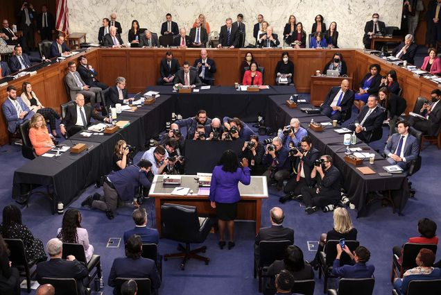 Photo of the Senate Confirmation Hearing For Supreme Court Nominee Ketanji Brown Jackson. Jackson, now an associate justice of the US Supreme Court (center), wears a purple blazer and black pencil skirt, and raises her right hand. A group of photographers sit before her and try to get a shot. Senators sit at tables in a U shape.