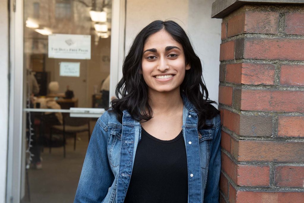 Portrait of The Free Press e-board’s Madhri Yehiya (CAS’24). She smiles, wears a jean jacket and black shirt and has olive skin, dark hair that reaches her shoulder. She lean against a brick wall outside a door that has a small daily free press sign on it.