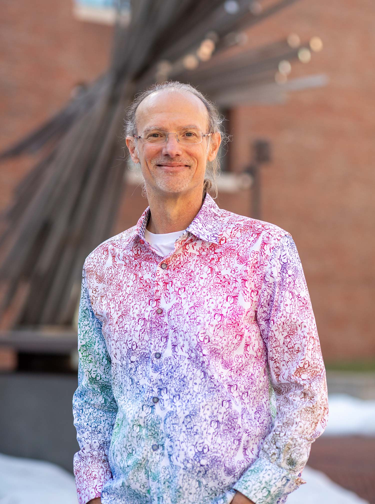 Photo of Mark Grinstaff, a middle-aged white man with glasses who wears a button-down shirt with a gradient, squiggly line pattern that fades from green and blue to red and pink. He smiles as he stands outside with his hands in his pockets.