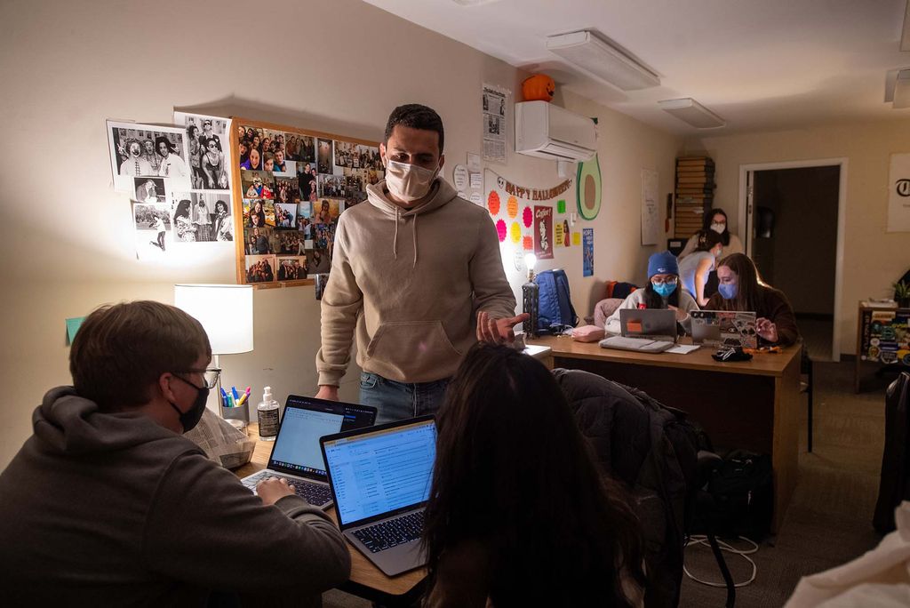 Photo of Editor-in-chief Jean-Paul Azzopardi (COM’23). wearing a tan sweatshirt and white face mask, on Print Night February 24 at The Daily Free Press office beneath Insomnia Cookies on Comm Ave. A few students on laptop sit in the foreground and speak with Jesus. A cork board on the wall at left is covered with photos.