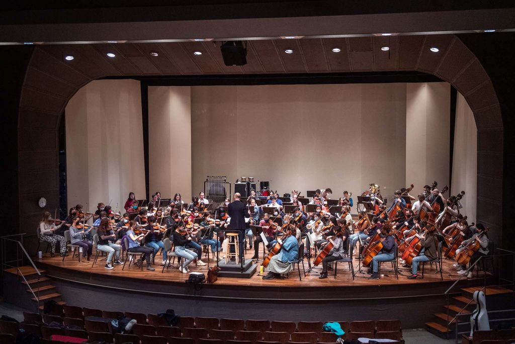 Wide shot of the BU orchestra practicing in the Tsai Performance Center at the end of March. Students play masked on stage.
