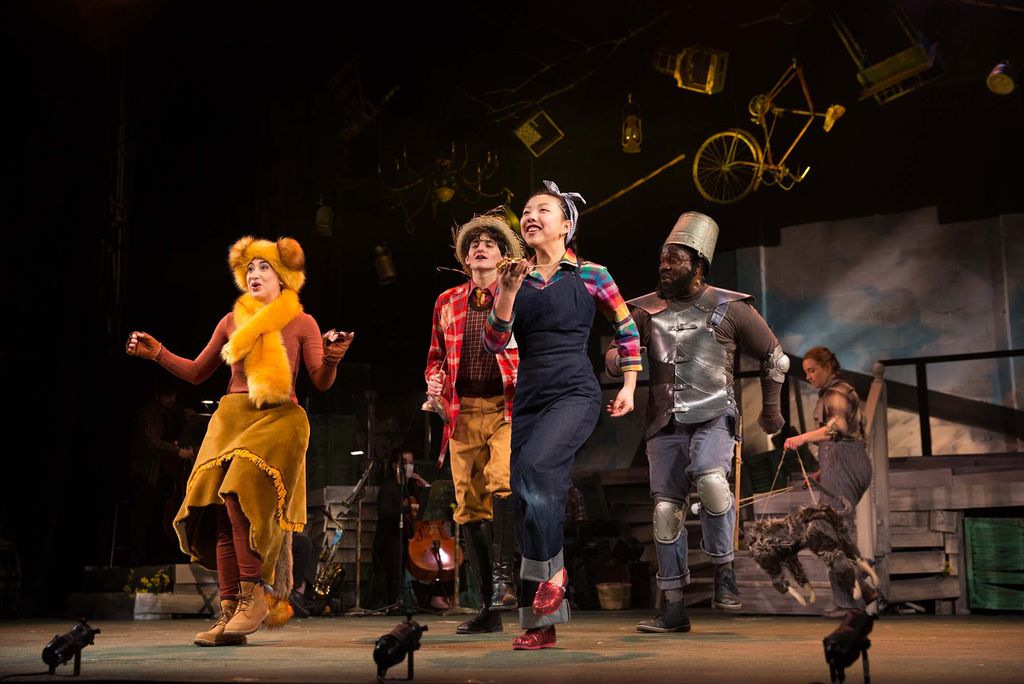 Photo, from left to right, of Krystale Hernandez as Lion, Max Seeling as Scarecrow, Lily Park as Dorothy and David Jiles as Tin Man, in Boston University’s production of “The Wizard of Oz,” during a dress rehearsal. The lion wears a furry hat with ears and fuzzy scarf, the scarecrow wears a plaid red shirt, orange pants, boots and a straw hat, dorothy wears overalls, red slippers, and handkerchief tied around her head, and the tin man wears a bucket on his head and a metal vest. The cast is a diverse group of young people.