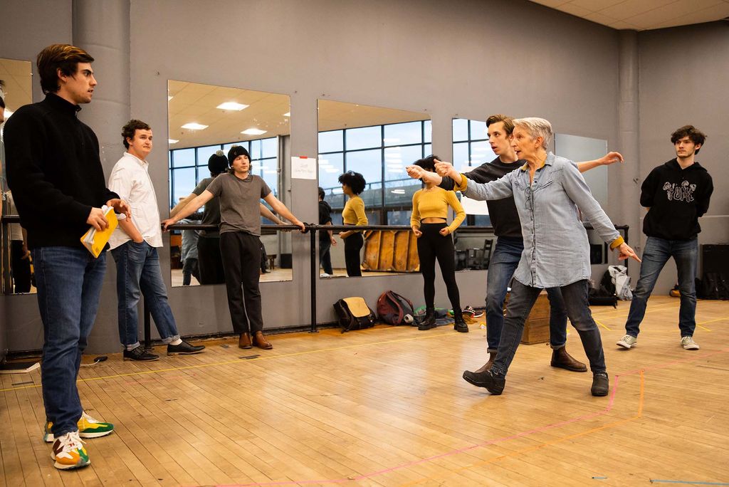 Photo of Judy Braha (CFA’08) working on what appears to be a sword fight scene with a student during a rehearsal for Shakespeare in Love. Braha wears a long, light blue button-down and black jeans. She's a white woman with short, gray hair. The student wears a black t and jeans. Both Braha and the student extend their arms forward as if holding a sword. Other students are seen looking on, some lean against the mirrored walls.