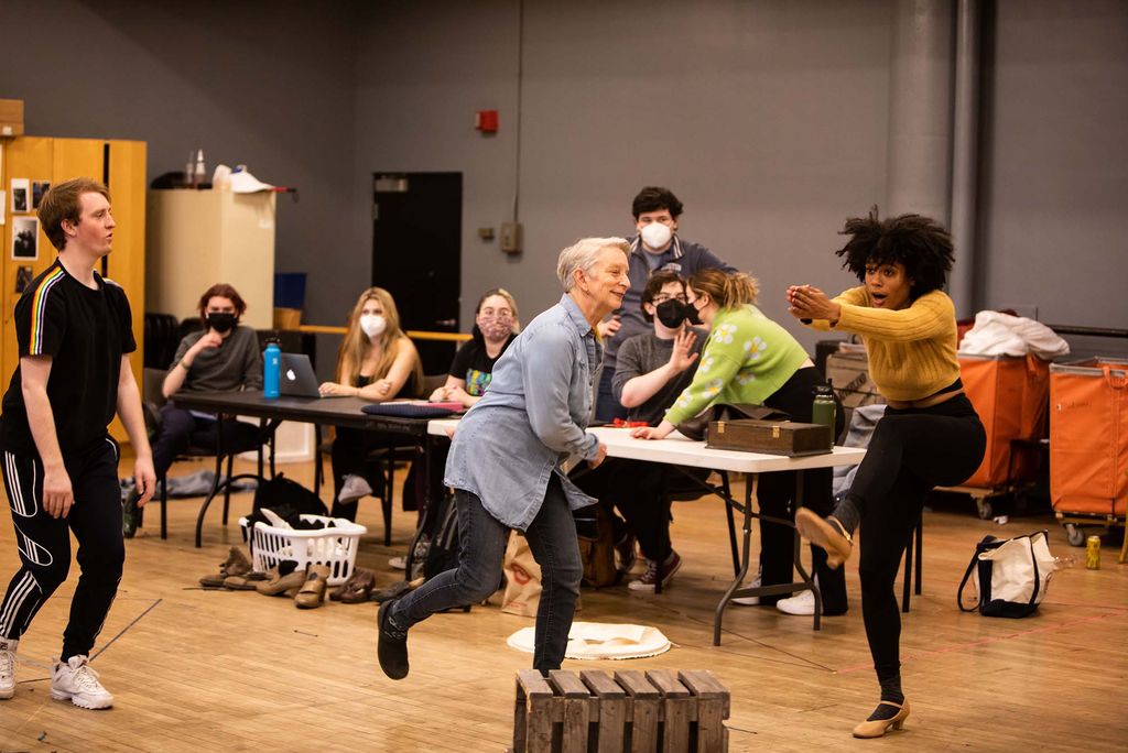 Photo of Judy Braha (CFA’08) working on a dance with a young student of color during a rehearsal for Shakespeare in Love. Braha wears a long, light blue button-down and black jeans. She's a white woman with short, gray hair. The student wears a mustard-colored sweater and black jeans. Both Braha and the student extend are seen mid dance move. Students sit at a table behind them.