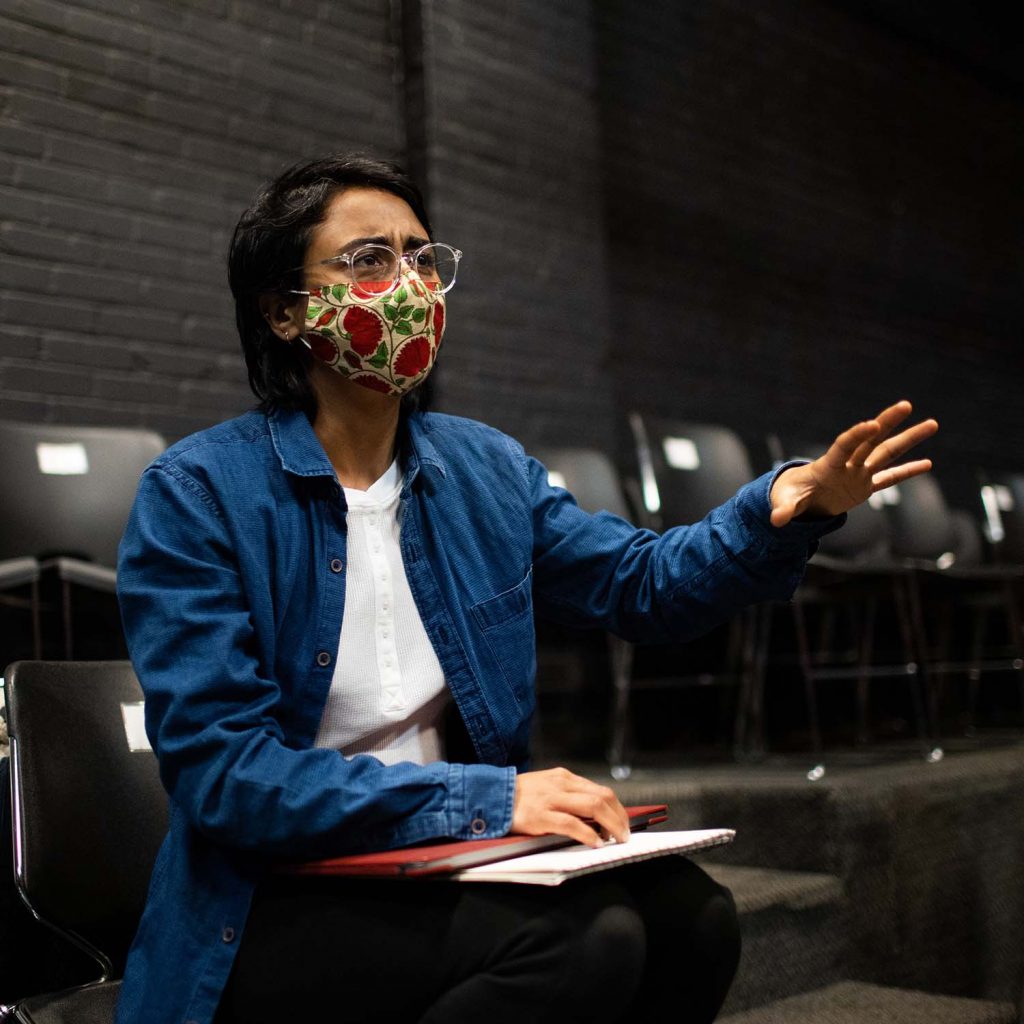 Photo of Fatima A. Maan, a graduate student from the School of Arts and Sciences who is an MFA playwright class of 2023. Mann wears round, clear-framed glasses, a long jean button down, a colorful floral face mask, and sits on a black chair as she directs the actresses in her play. She looks engaged and raises her left hand toward the stage during the rehearsal.