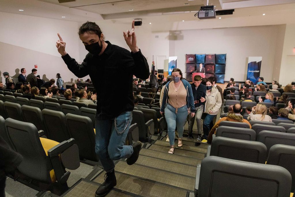 Photo of Diarmuid Ryan (CAS'23), left front,  and other students walk out in protest on Michael Knowles' talk on "Teaching Kids Sex and Gender Ideology," with BU YAF at the Law Auditorium on Thursday. Ryan wears a black shirt, black mask, jeans, a pocket chain and boots, and walks out while flipping off Knowles. Other students walk up the auditorium stairs behind Ryan and Knowles can be seen at the podium in the background. 