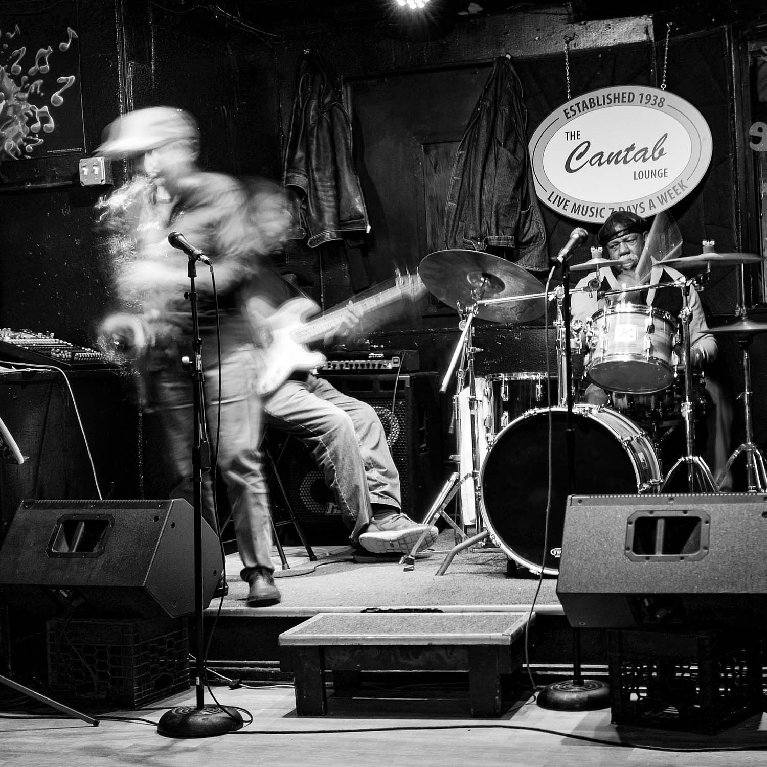 Black and white photo of a band performing at the cantab lounge. The photo was taken in such a way that the band members are seen in motion. A drummer, lead singer and bass player sitting in the back are seen performing.