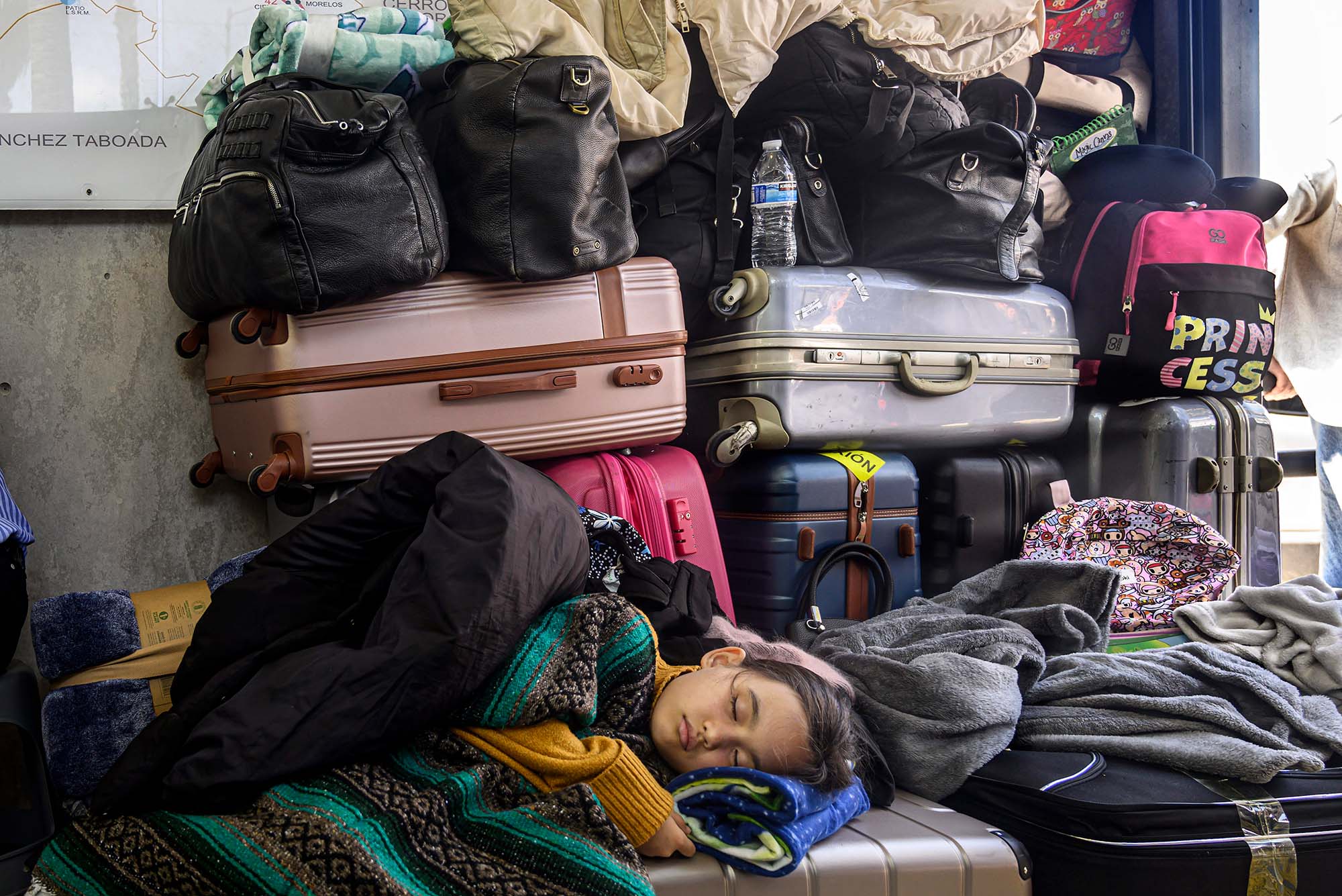 Photo of a young Ukrainian girl sleeping on top of a pile of suitcases. She is sleeping underneath a few thick blankets and a large pile of suitcases is seen behind her.