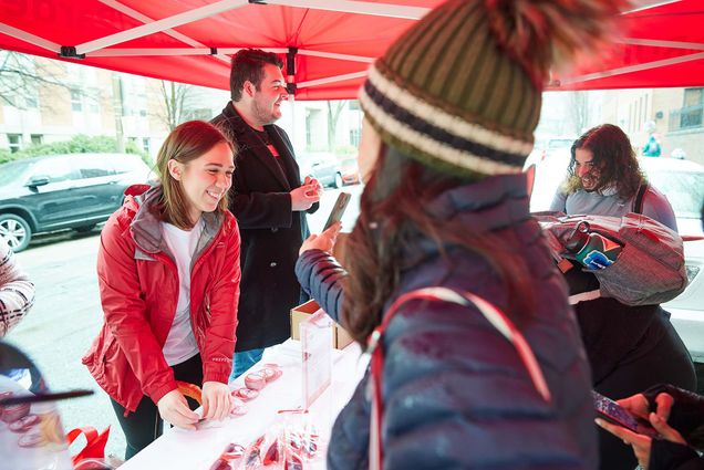 Photo of a person in a green winter hat and poofy jacket showing their phone to a young white woman with dirty blonde hair wearing a red jacket. She stands behind a table next to white man in a black rip up.