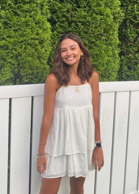 Portrait of Hannah Zobair (CAS’22). She is a white woman with shoulder length dirty blonde hair. She smiles and wears a white dress and stands in front of a white fence with hedges behind her.