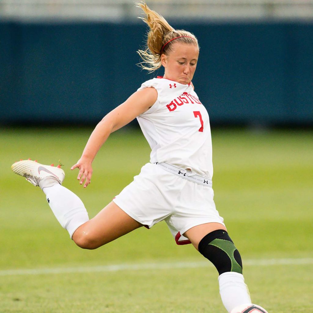 Action photo of BU women's soccer player, Ann Marie Jaworski. She is caught poised ready to kick the ball and wears a white "Boston" jersey, shorts, shin guards, and cleats. She also wears a black knee brace.