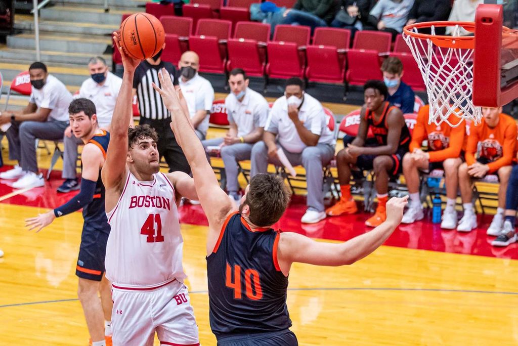 Action photo of BU men's basketball player, Sukhmail Mathon. A young man with a short curly brown hair is shown taking a shot for the hoop. An opposing player in a blue and orange jersey jumps in front of him with arms raised as another opposing player in a similar uniform looks on behind them. Various coaches and player in white BU shirts, as well as the opposing team in blue jerseys can be seen sitting at the edge of the court in the distance.