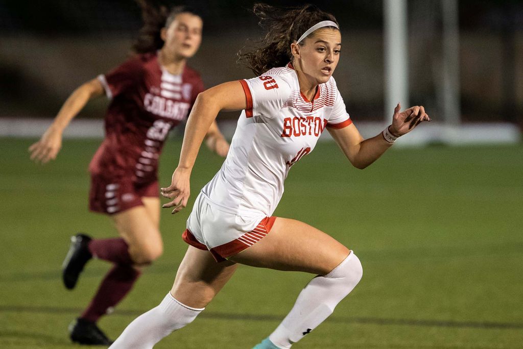 Photo of BU women's soccer player Abigail McNulty. A young woman with long, curly brown hair tied back is shown mid-run on a soccer field. She wears a white and red "Boston" soccer uniform. A Colgate athlete wearing a burgundy soccer uniform is shown running behind her in the blurry background.