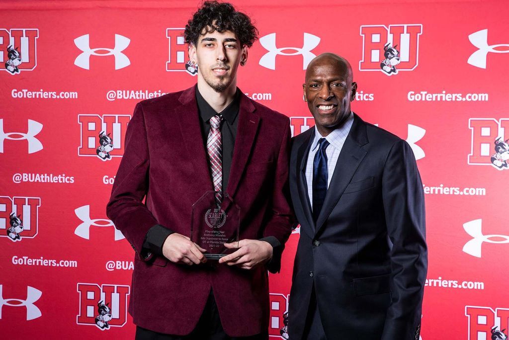 Photo of Anthony Morales, a BU men’s basketball player. He stands with a small clear trophy held in front of him, next to his coach, Joe Jones, a middle-aged Black man wearing a black suit ensemble and velvet blue tie. They both stand in front of a red backdrop with the BU school logo, the Under Armour logo, and the words "@BUAthletics" in an intermittent pattern.