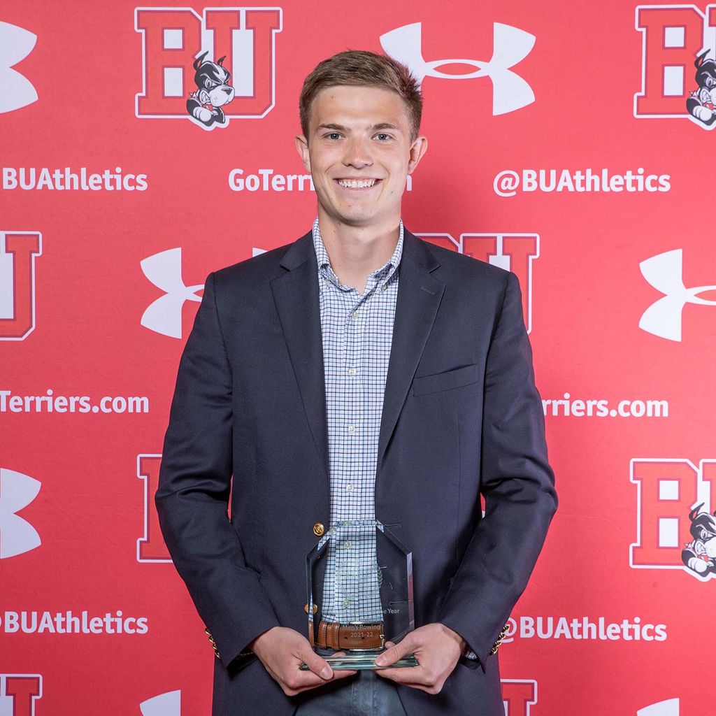 Photo of BU men's rowing athlete Harrison Steck. Steck is shown wearing a light blue and white plaid collared shirt, navy blazer, and dark grey pants as he siles and poses with a small, clear trophy held with both hands in front of him. He stands in front of a red backdrop with the BU school logo, the Under Armour logo, and the words "@BUAthletics" in an intermittent pattern.