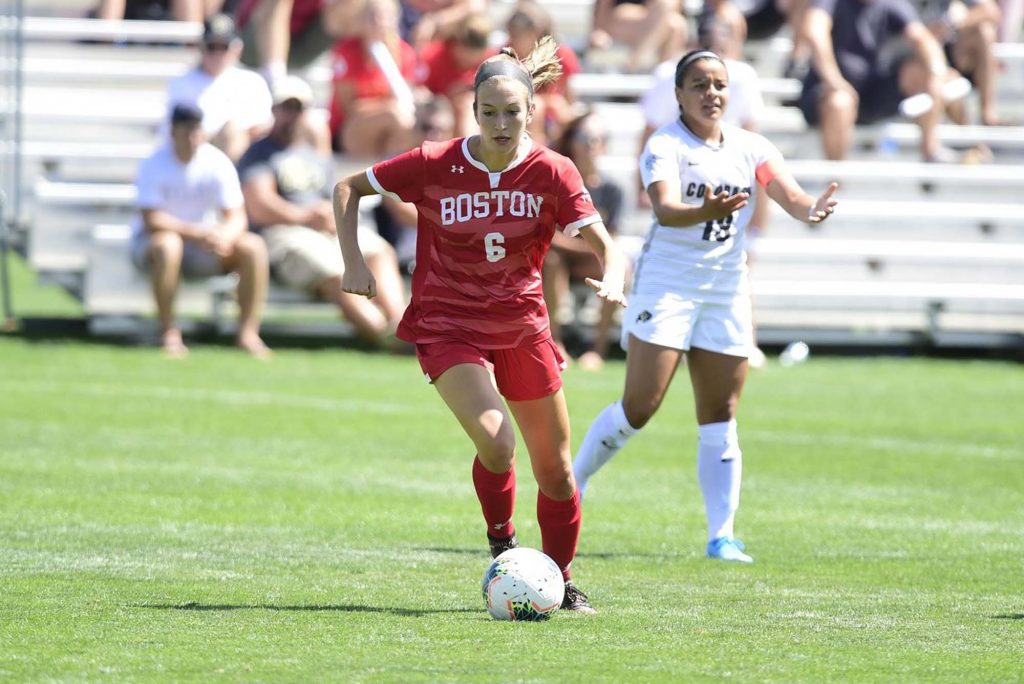 Photo of BU women's soccer player Sophia Woodland. She is caught mid-game as she runs with the ball down field. She wears a red Boston jersey, shorts, and shin guards as she moves away from another opposing player in white.