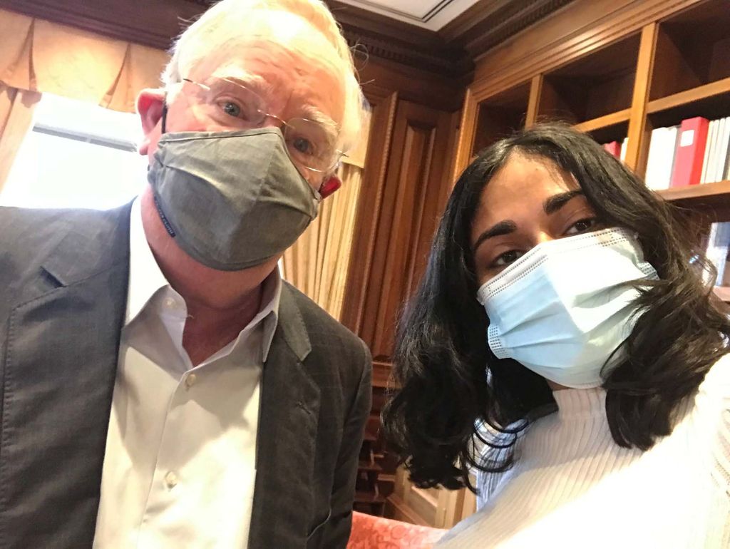 Photo of Madhri Yehiya (CAS’24), a young brown woman with short dark hair wearing a facemask, taking a selfie with BU President Bob Brown, an older white man who wears glasses and a gray face mask. In the background, the president's wood-paneled office is seen.