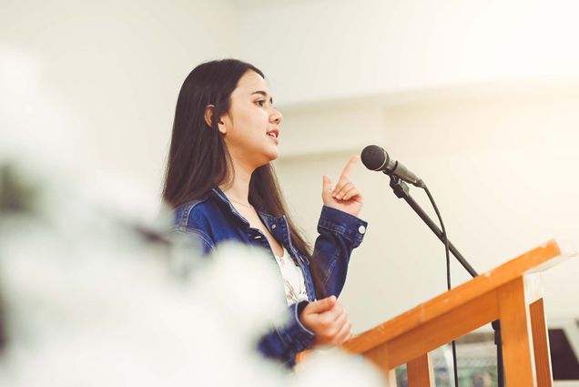 Photo of a young woman, either a high school or college student with brown hair, wearing a jean jacket, speaking at a wooden podium into a microphone. She gestures with her left hand and appears to be mid-speech.