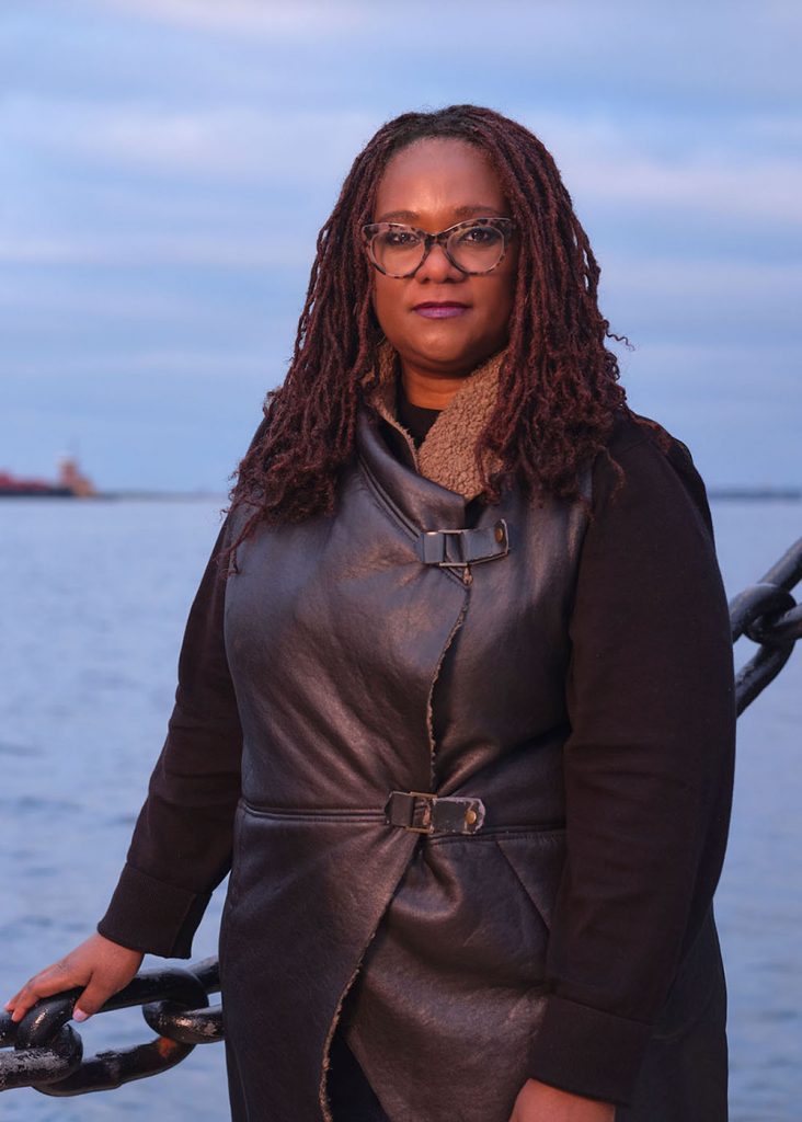 Photo of Deborah Douglas along boston harbor. She wears a black leather jacket, has shoulder-length, crimson locs, and rests her hand on a large middle chain fence. Behind, the harbor at twilight is seen.