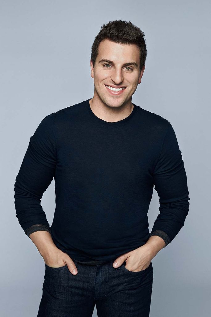 Photo of Brian Chesky, Airbnb CEO, who wears a black long-sleeve shirt, and smiles, his hands in his jean pockets on a neutral gray background. He is a white man with short brown hair.
