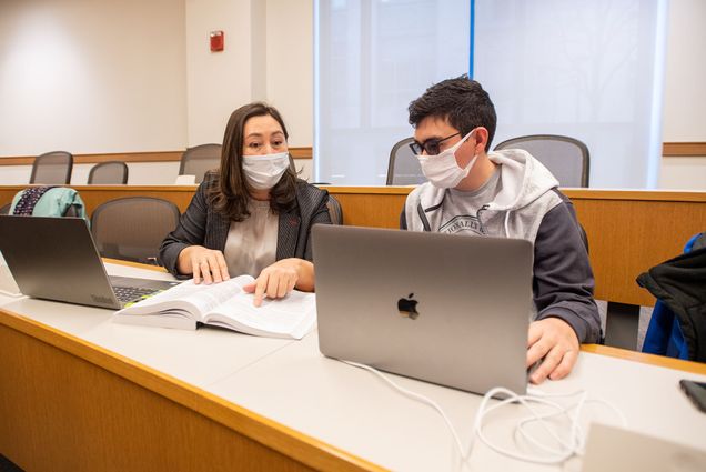 Photo of Gulfiya Nassyrova (LAW’22), left, and her son Nariman Temirbekov (LAW’22) beginning to study for a course on security transactions March 3 at LAW. Gulfiya is a middle-aged woman with light skin and shoulder-length brown hair and wears a face mask. She points to a line in a text book and has her laptop open in front of her. Nariman is a young man with short dark brown hair and transition glasses. He wears a gray sweatshirt and sits in front of a laptop.