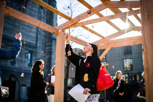 Photo of Alex Barnes (CAS 24, a young person with long brown hair and an undercut, who wears a black jacket with the BU Logo, interacting with one of the objects hanging from a wooden structure. The structure is part of a student public art project titled "Home" by Kabita Das (CFA'20) on Monday, April 4, 2022.
