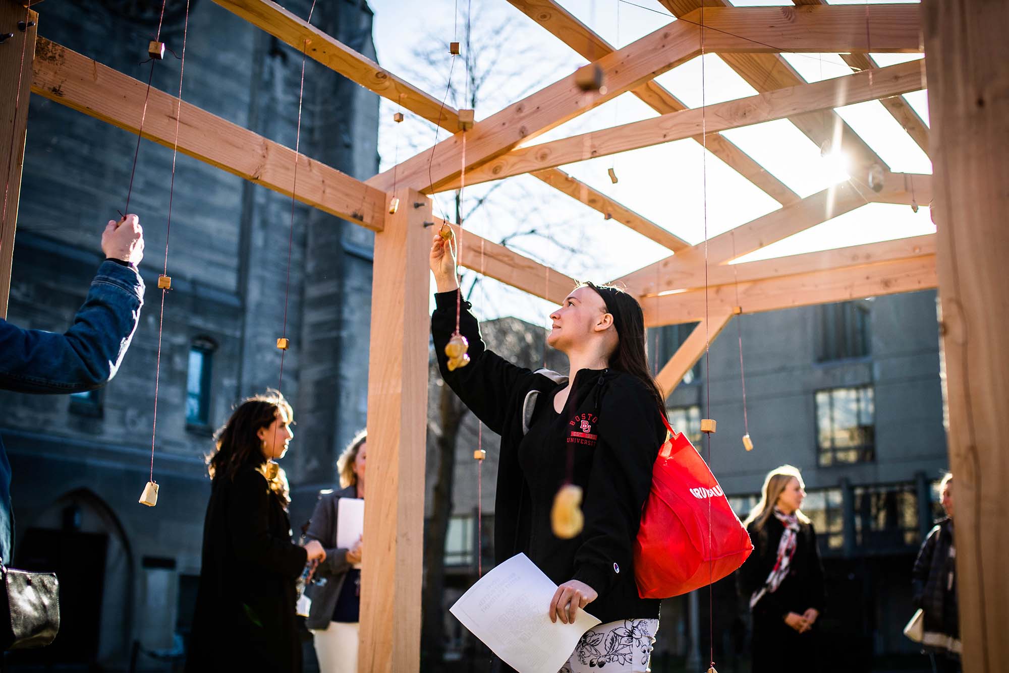 Photo of Alex Barnes (CAS 24, a young person with long brown hair and an undercut, who wears a black jacket with the BU Logo, interacting with one of the objects hanging from a wooden structure. The structure is part of a student public art project titled "Home" by Kabita Das (CFA'20) on Monday, April 4, 2022.
