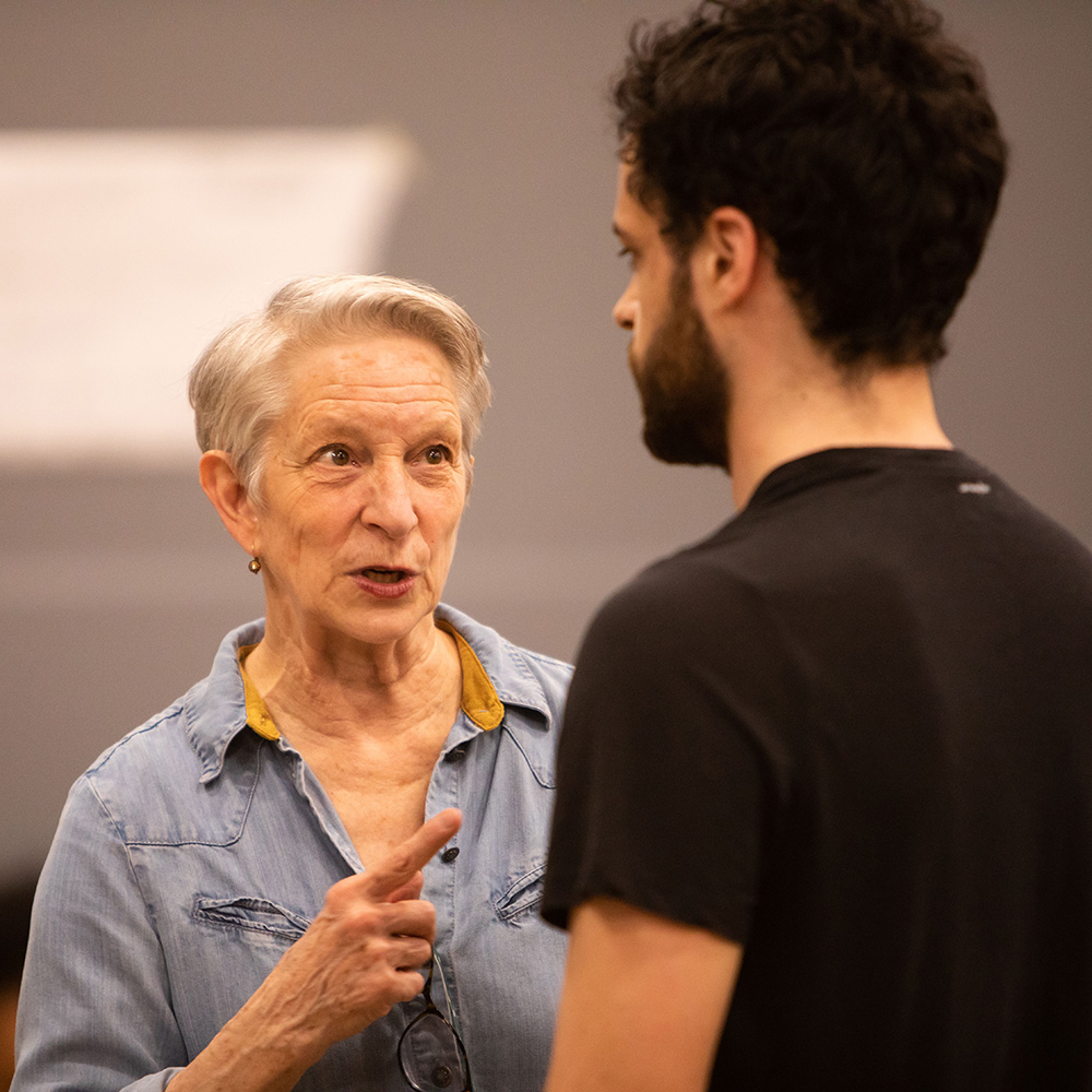 Photo of Judy Braha (CFA’08) speaking to a student during a rehearsal for Shakespeare in Love. Teh student has short brown hair and a beard, and wears a black t-shirt. Judy points at the student with her hand and appears to be giving directions. 