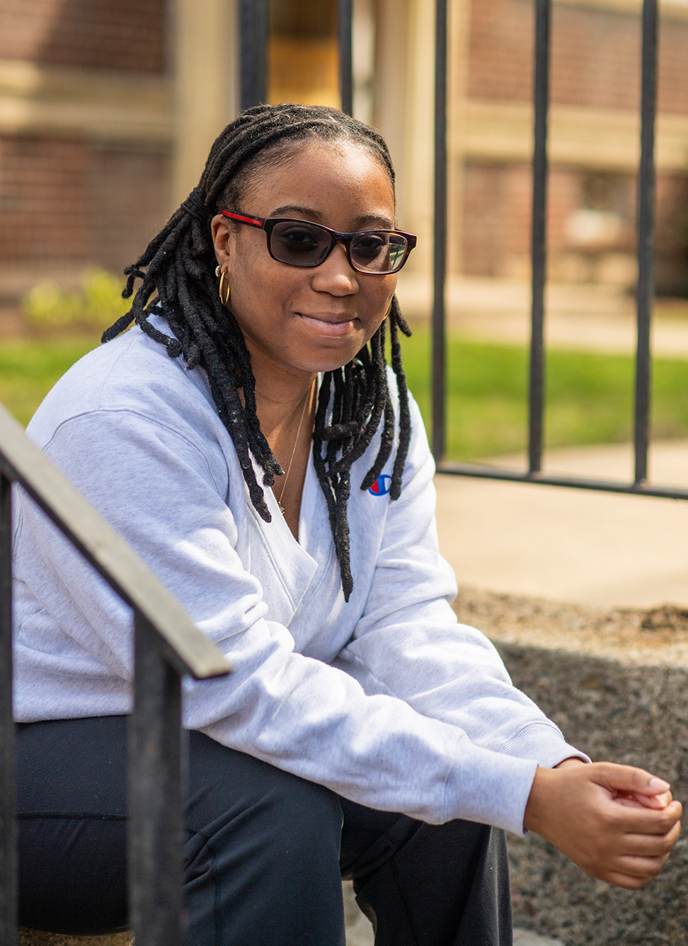 Photo of Keanna Goddard, a young black woman with long, black dread locs and wearing tinted glasses sits with hands folded in prayer on cement stoop steps on a bright day.