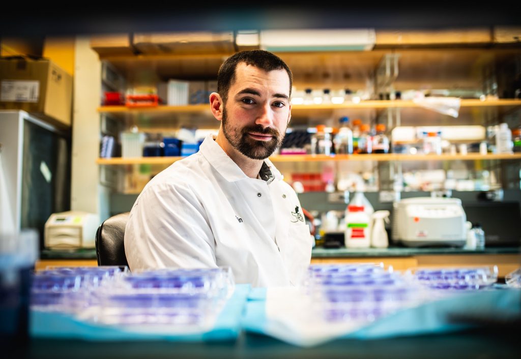 Photo of Devin Kenney, a PhD student, as he poses for a photo at the National Emerging Infectious Diseases Laboratories. Kenney looks to the right towards the camera overtop a set of test tubes with light purple tops. Blurry lab shelves can be seen in the background.