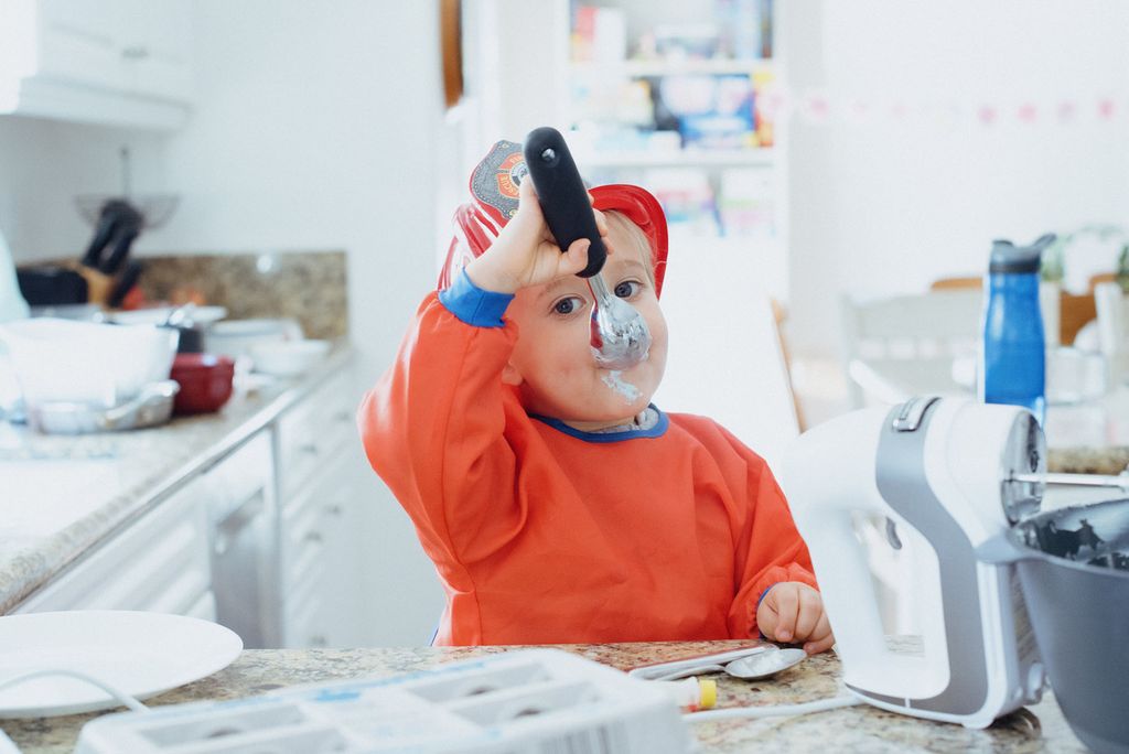 Photo of a toddler wearinf an orange sweatshirt and plastic fireman hat messily eating ice cream from an ice cream scoop. 