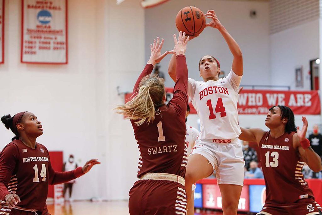 Photo of a BU Women’s basketball taking a jump shot for the basket. She wears a white and red "Boston" jersey and is surrounded by three Boston College players trying to defend. One jumps in front of her with arms raised overhead, one is behind her defending against an unseen BU athlete, and one stands off to the left as she looks on. all three wear burgundy red basketball uniforms that read "Boston College".