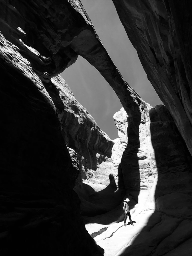 Black and white photo of a person standing in the Arches National Park. They stand under a large rock cliff formation. The small size of the person is juxtaposed with the large formations.