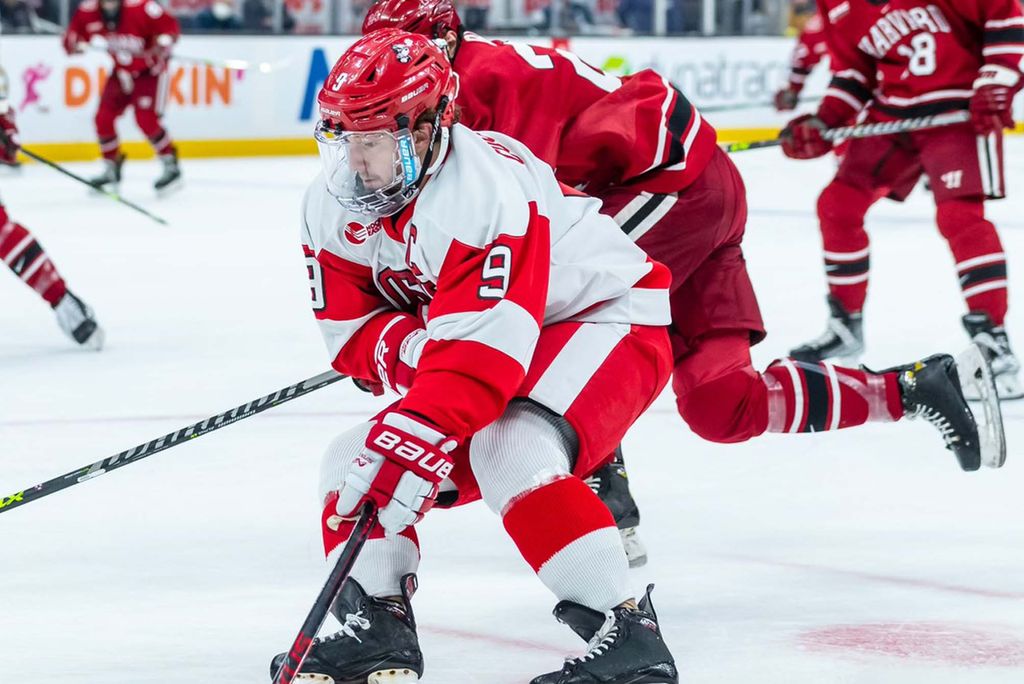 Photo of men’s ice hockey captain Logan Cockerill. Cockerill is shown mid-game as he skates with the puck down the ice away from an opposing Harvard player. He wears a red and white BU ice hockey uniform and black skates. Opposing Harvard players wearing crimson hockey uniforms skate around him.