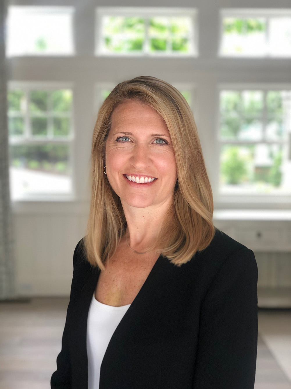 Headshot photo of Constance Noonan Hadley. A white woman with blonde hair wearing a plain white shirt and black blazer smiles in a spacious office.