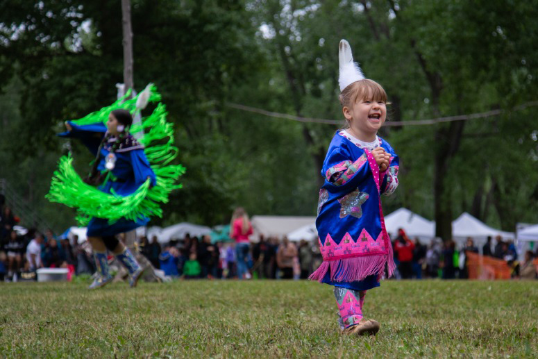 Photo of a small child wearing a blue Native outfit as she runs down a field with hands clasped. A woman wearing another blue Native outfit with large neon green frills dances in the background