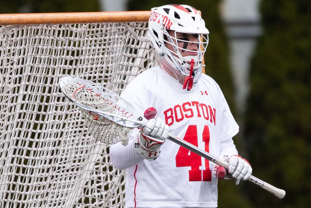 Photo of Matt Garber, a a young white man who wears a white jersey, number 41, and wears a lacrosse helmet. He holds a large lacrosse stick and is seen in front of the net.