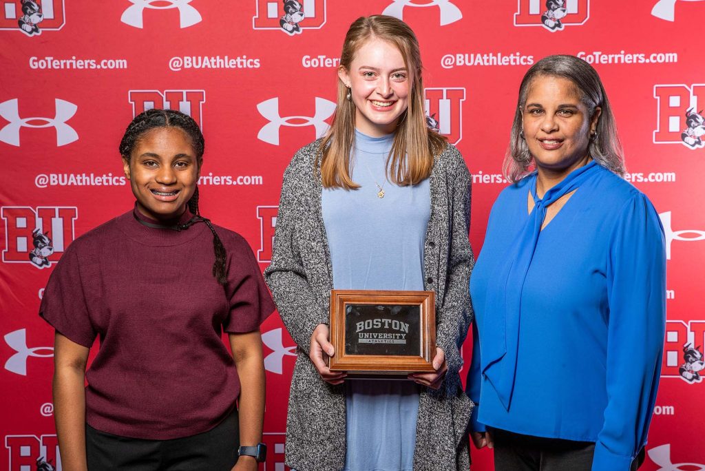 Photo of Kylie Graves, a young white woman with strawberry blonde hair, posing with her award and the family of Paul Lewis. At left, a young woman of color is seen, and at right, an older woman of color in a blue blouse.