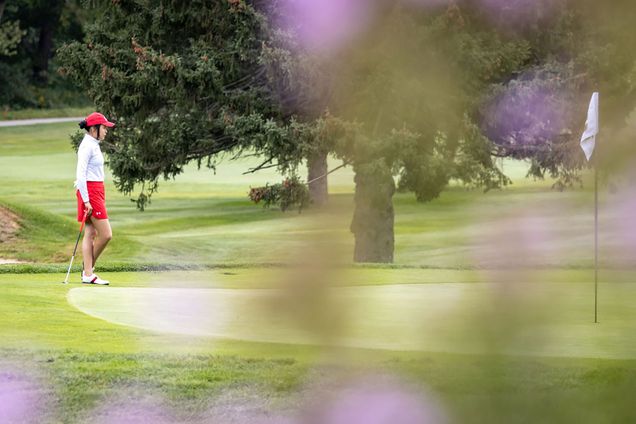 Photo of senior Hanako Kawasaki (Questrom’22, SHA’22), taken from a distance during a golfing match. Kawasaki stands leaning slightly on her club at the edge of a green, she wears a white long sleeve, red skirt and red hat. In the foreground, a tree branch blurs the right half of the photo.