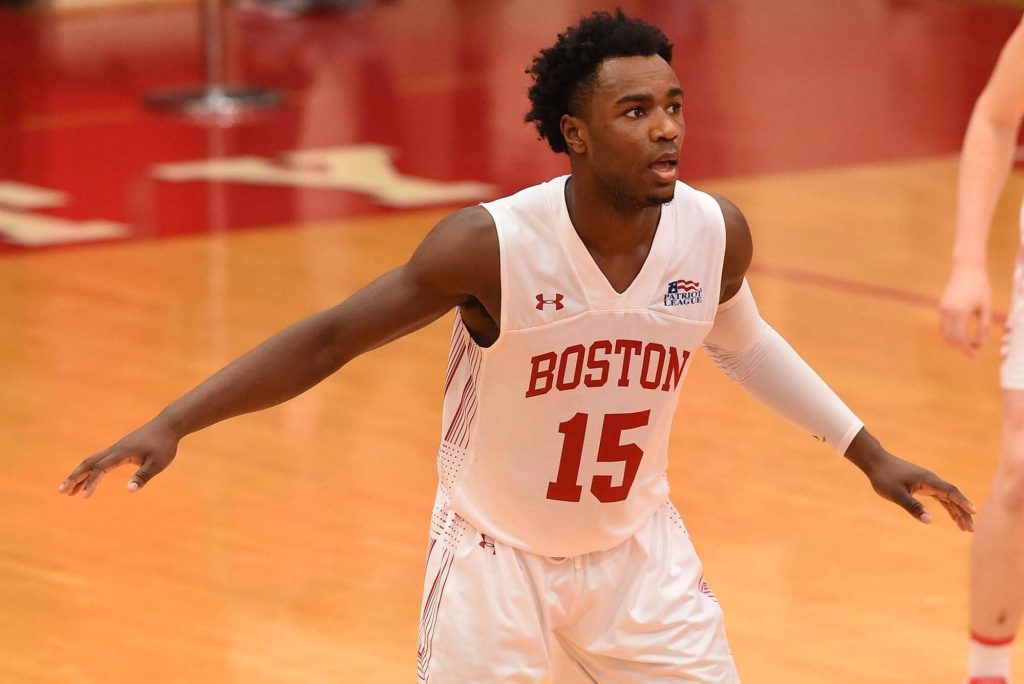 Action photo of BU men's basketball player, Jonas Harper. A young black man with a short afro is shown arms out in a guarding stance on a basketball court. He wears a white Boston jersey and white shorts.